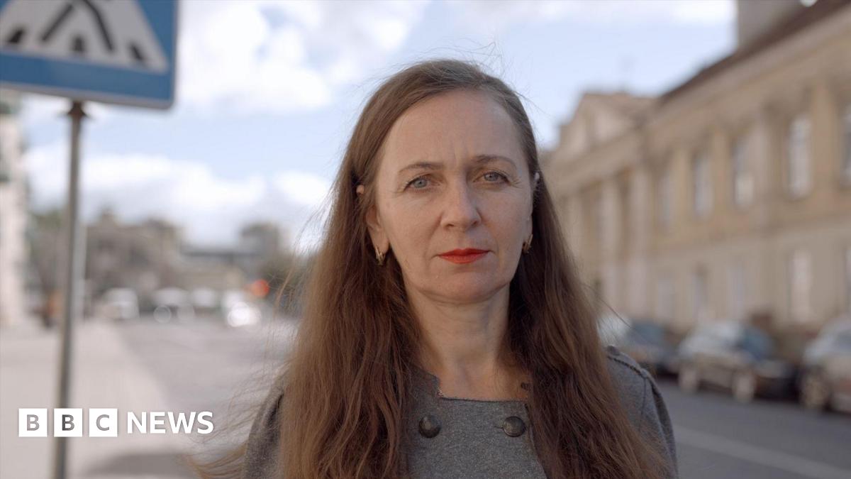 A woman with long brown hair stands in a street and stares at a camera