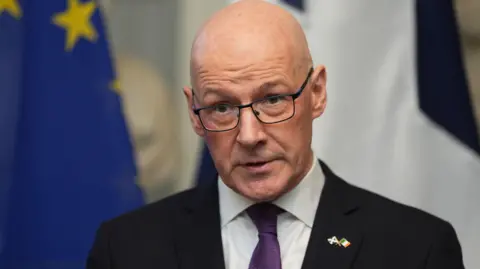 PA Media John Swinney stands in front of an EU flag wearing a black blazer, a white shirt and a purple tie