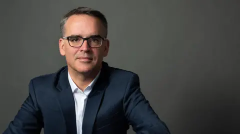 Stephanie Bonnas Weber has short browny-grey hair and wears black rimmed glasses. It's a studio shot and he's wearing a blue suit