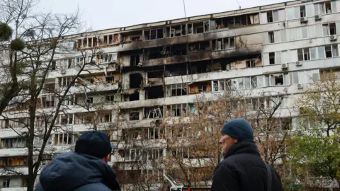 EPA Two men with their backs to the camera, looking at a residential building which stands in front of them. the top centre of it is blackened and damaged following a strike
