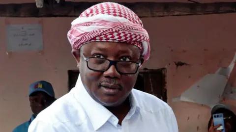 Reuters Umaro Sissoco Embaló in a white shirt and checked red and white head scarf looks at the camera as he votes on 23 November 2025.