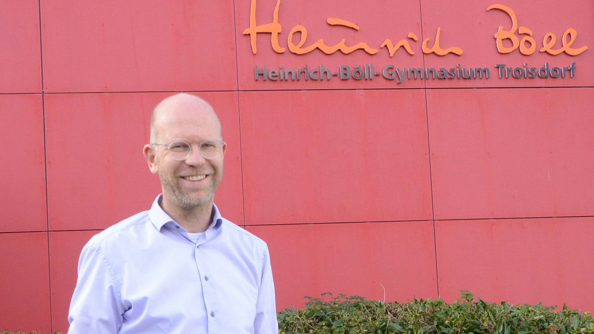 Ein Mann vor einer roten Hauswand mit der Aufschrift Heinrich-Böll-Gymnasium, Troisdorf