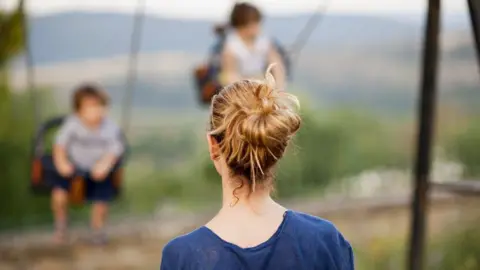Getty Images Stock image of a woman with her back to the camera looking at two children on swings.
