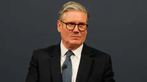 Reuters Sir Keir Starmer has grey hair which is slicked back to one side of his head with shorter sides and is wearing oval glasses, a black suit jacket, white shirt and blue tie with yellow triangular print on it. He has a microphone attached to his tie and is looking to the side with a serious expression. The background is dark grey.