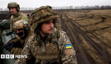 Ukrainian soldiers in full battle dress sit on a cannon in a muddy field.