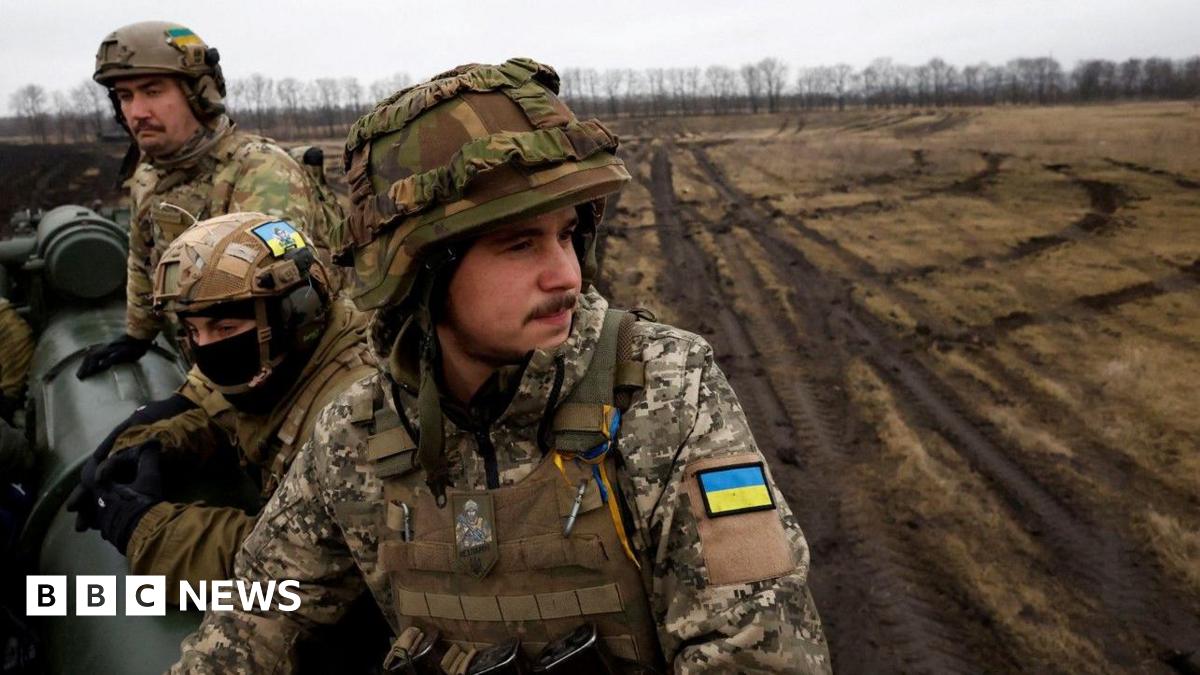 Ukrainian soldiers in full battle dress sit on a cannon in a muddy field.