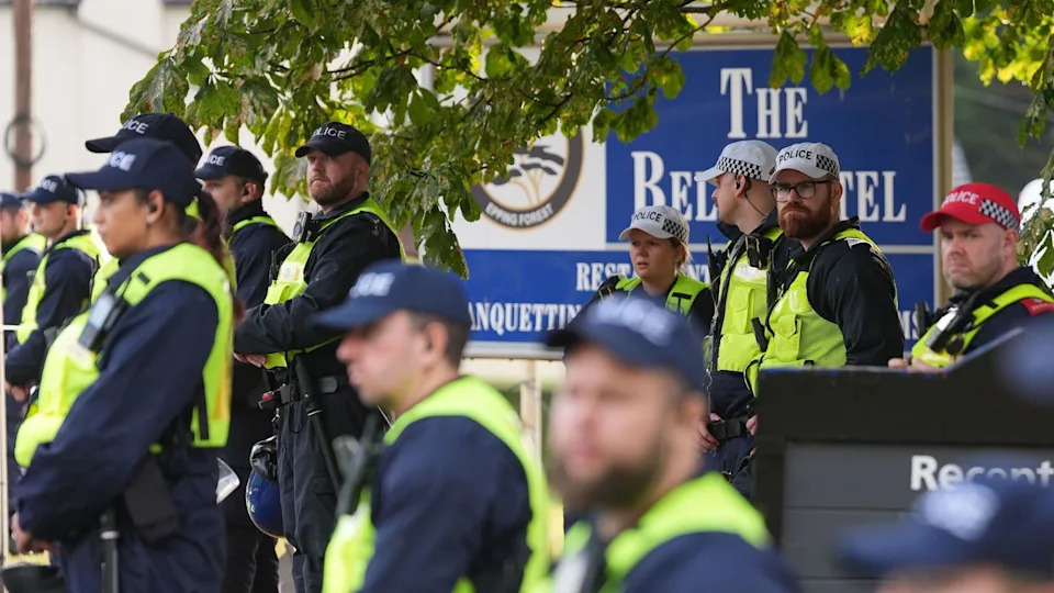 A large group of police officers, all wearing caps and hi-vis vests, standing in front of a large blue sign outside The Bell Hotel, which advertises its name.