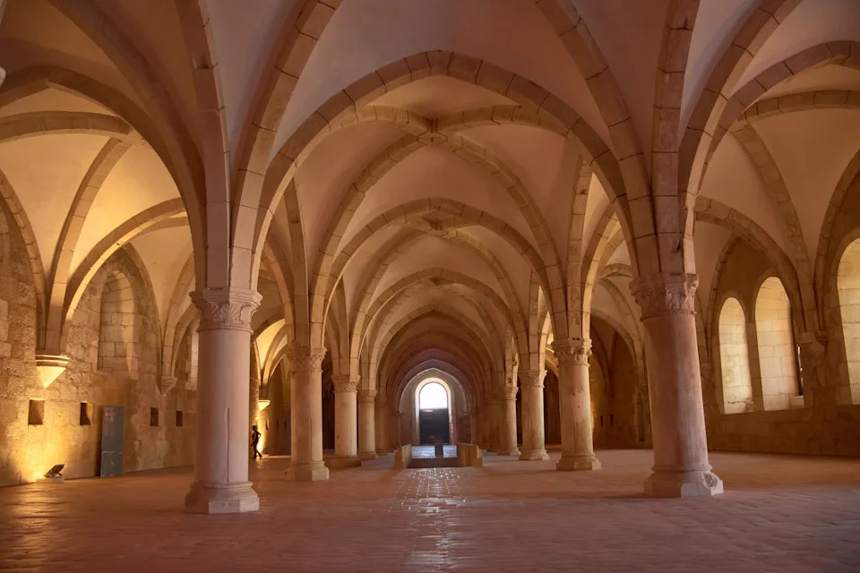 The novices' dormitory at the Alcobaca Monastery extends like a large Gothic hall with stone columns, a soaring vault, and natural light pouring in through arched windows. The symmetry and silence of the space evoke monastic life and medieval spirituality. Ideal for representing sacred architecture, historical heritage, and cultural tourism in Portugal