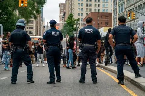 Reuters Four police officers have their backs to us as they provide security in Union Square as popular live streamer, not shown, stages a giveaway. They have NYPD on their shirts and one of them wears a helmet. 