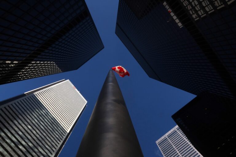 A Canadian flag flies in front of a tall black pole, surrounded by towering buildings.