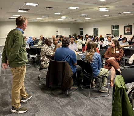A group of people sit in a room around tables with one person standing and looking at them