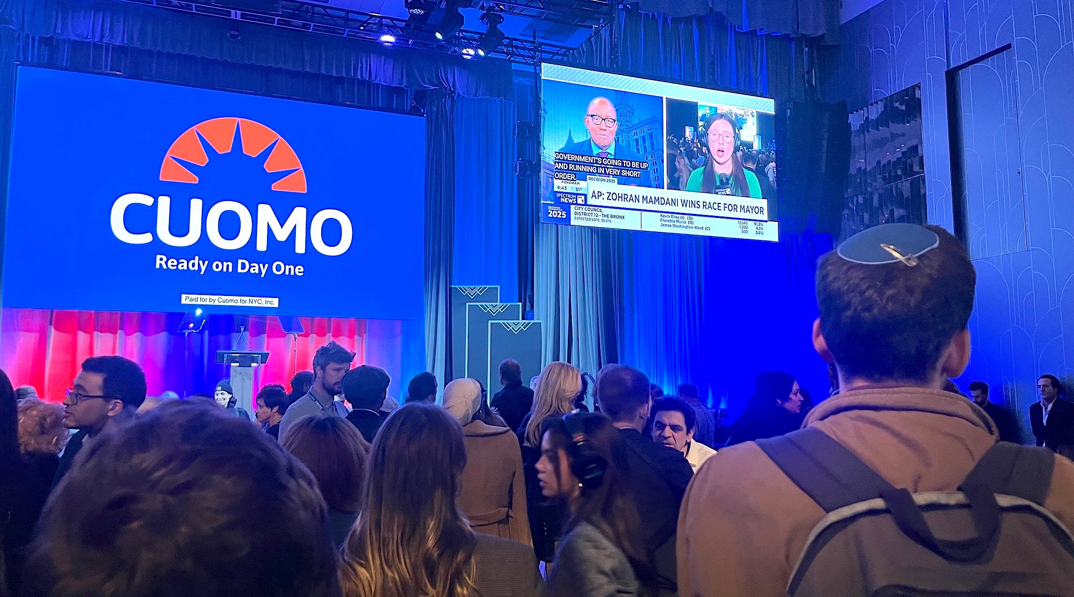 A photo of a man in a kippah watching the election be. called for mamdani on a projector in a crowd.