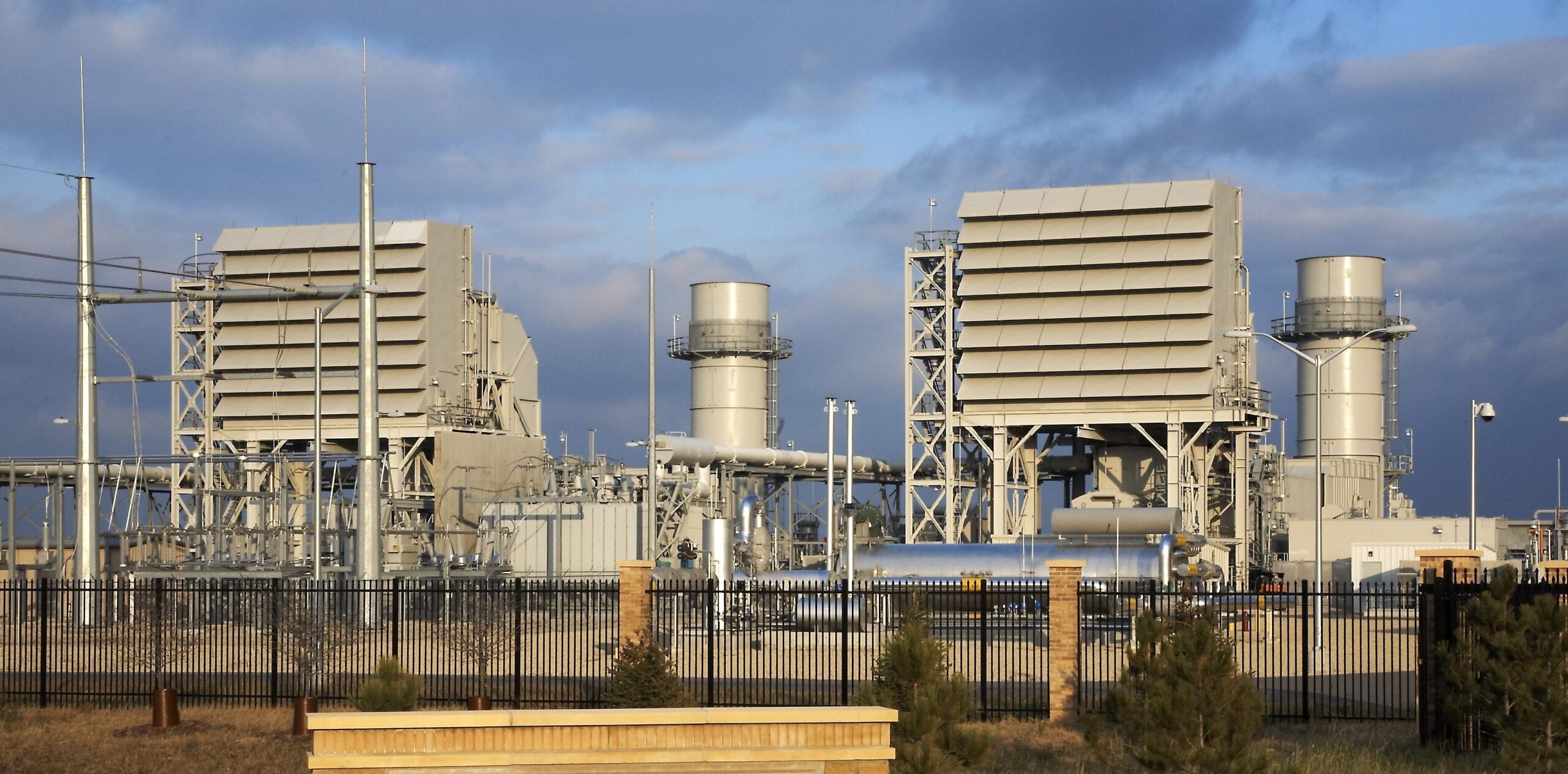 A natural gas power plant with three large turbine generator units, surrounded by industrial piping and fencing, under a cloudy sky.