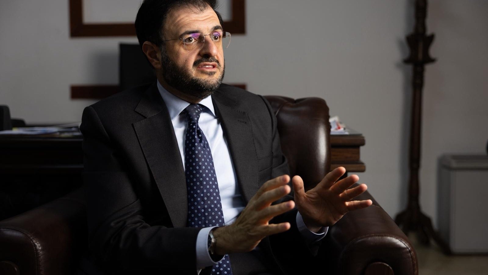 A man in a suit sits in a leather chair, gesturing with his hands while speaking, in a well-lit office setting.