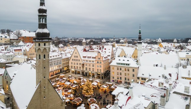 An aerial view over Christmas Market stalls in Tallinn, Estonia, surrounded by snow-topped buildings, on a cloudy day.