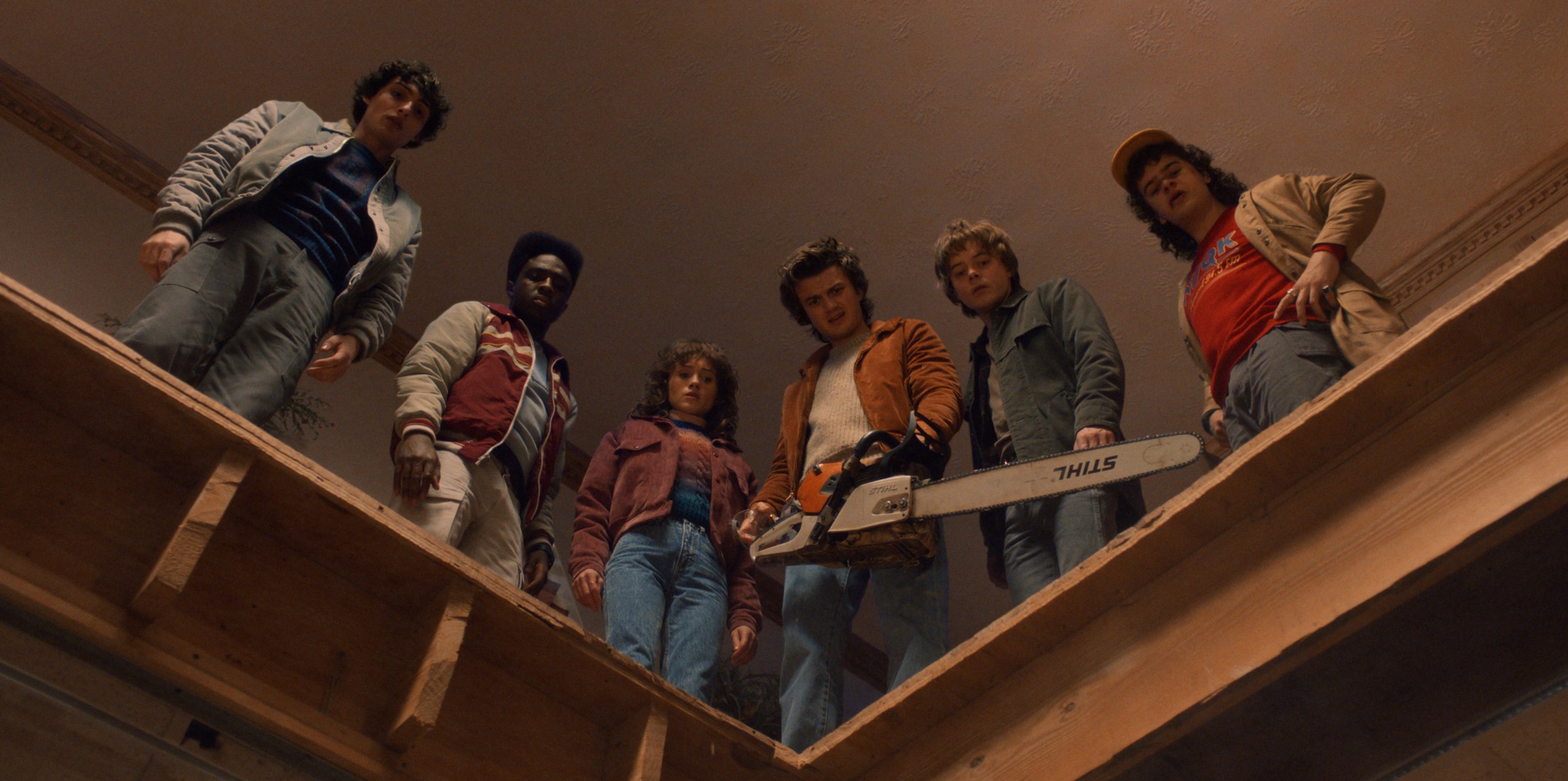 Six teenagers standing together on a ledge within a building. The teens are all looking down at something in confusion.