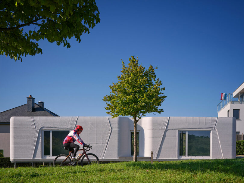 A cyclist rides past a modern white 3D-printed house with large windows and a tree in the front yard under a clear blue sky.