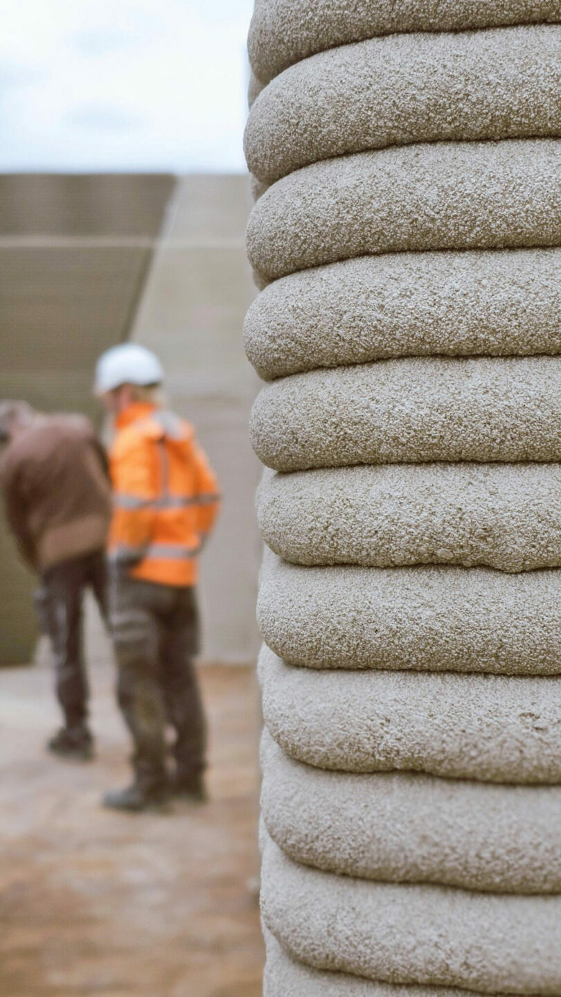 Close-up of layered concrete structure in foreground with two people, one in a high-visibility vest and helmet, standing and talking in the background on a construction site.