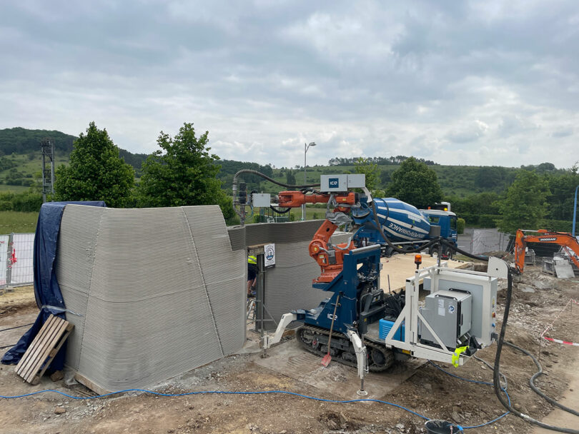 A robotic 3D printer constructs a concrete wall at an outdoor construction site, with machinery and trees visible in the background.