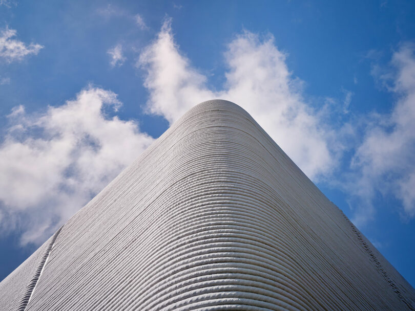 Low-angle view of a modern building with curved edges and horizontal lines against a blue sky with scattered clouds.
