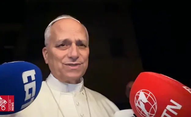 Person in clerical attire speaks into microphones from various news outlets