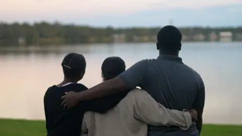 Liam Weir/BBC Marven, Rochelle and Guerline, pictured from behind, with their arms around each other looking out over a lake. Marwen is wearing a grey, collared T-shirt and is much taller than the two women.