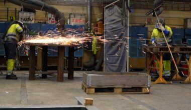 To personer arbeider i en verksted med sveising. Den ene sveiser ved et bord, og gnister flyr. Begge har på seg verneutstyr og refleksvester. Omgivelsene inkluderer verktøy og annet utstyr.