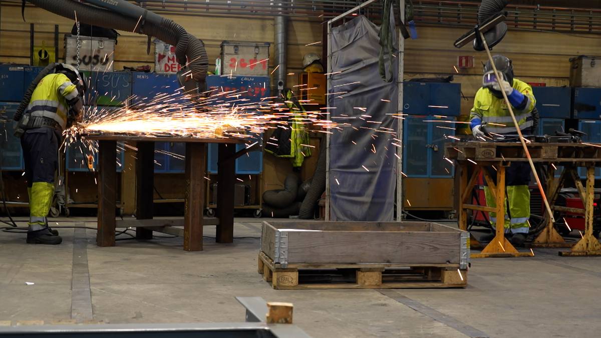 To personer arbeider i en verksted med sveising. Den ene sveiser ved et bord, og gnister flyr. Begge har på seg verneutstyr og refleksvester. Omgivelsene inkluderer verktøy og annet utstyr.