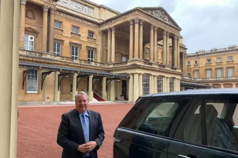 Facebook Jay Bloom, a middle-aged man with grey hair, wearing a navy suit and a blue shirt and tie, standing next to a dark coloured Range Rover in the quadrangle of Buckingham Palace, with its imposing portico behind him.