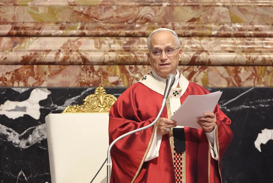A clergyman in ceremonial robes speaks at a podium inside a marble-walled hall, holding a document
