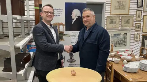 The Cotswold Auction House Two men - Niall Fry on the left who is wearing a white checked shirt and a grey blazer, and Marios Theocharous, who is wearing a dark shacket with a dark shirt beneath and has grey hair - shake hands as they stand in an auction house. In front of them, there is a small pine wooden table, with a small jug on it.