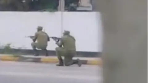 X Two men in green police uniforms kneeling down pointing rifles down a street