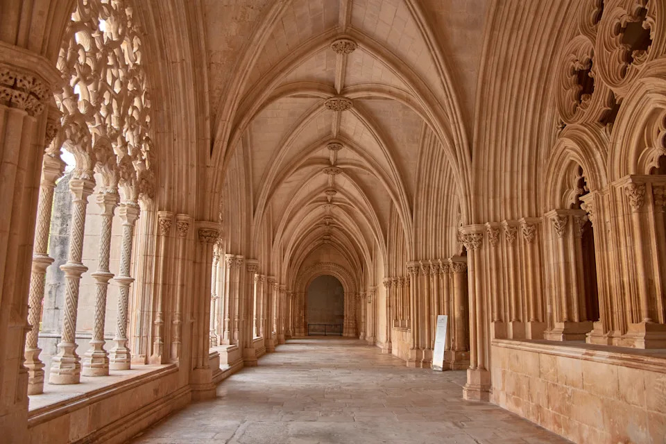 The cloister of the Batalha Monastery unfolds with Gothic elegance in a sequence of pointed arches, ribbed vaults, and carved columns that guide the eye to the background. Natural light passes through the lateral tracery, casting soft shadows that enhance the texture of the stone. Ideal for depicting medieval architecture, cultural heritage, and European spirituality