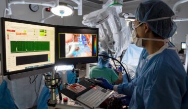 Surgeon look at computer screens and uses mouse inside operating room. A large surgical robot is seen in the background, alongside another surgeon.