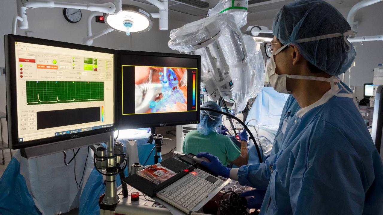 Surgeon look at computer screens and uses mouse inside operating room. A large surgical robot is seen in the background, alongside another surgeon.