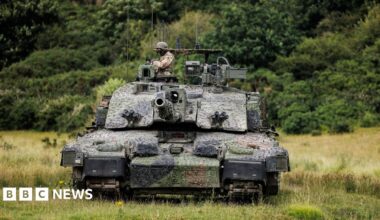 The Megatron variant of the Challenger 2 tank, heavily camouflaged, with a soldier wearing a helmet in the turret, advancing across a green field, with trees in the background