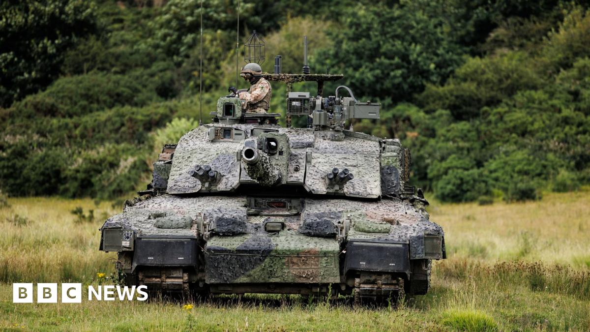 The Megatron variant of the Challenger 2 tank, heavily camouflaged, with a soldier wearing a helmet in the turret, advancing across a green field, with trees in the background