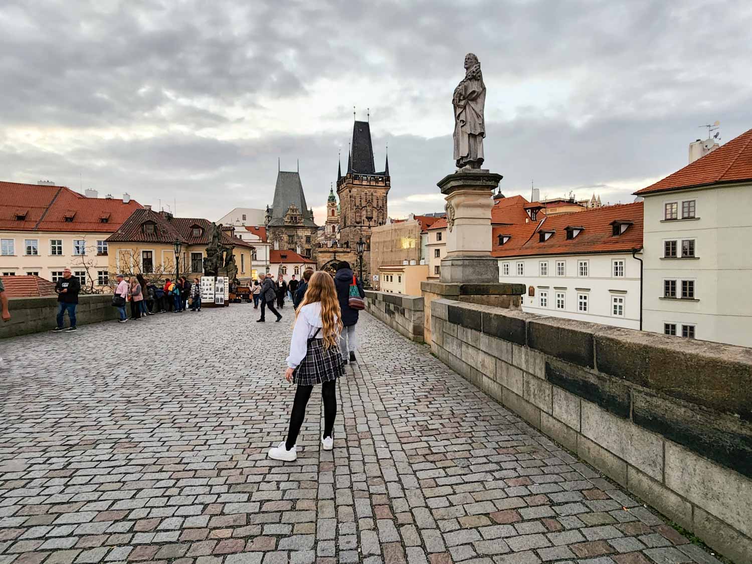 My daughter stands at the Mala Strana end of the Charles Bridge towards sunset with one of the statues visible and buildings beyond - one of the unmissable sights of Prague with kids