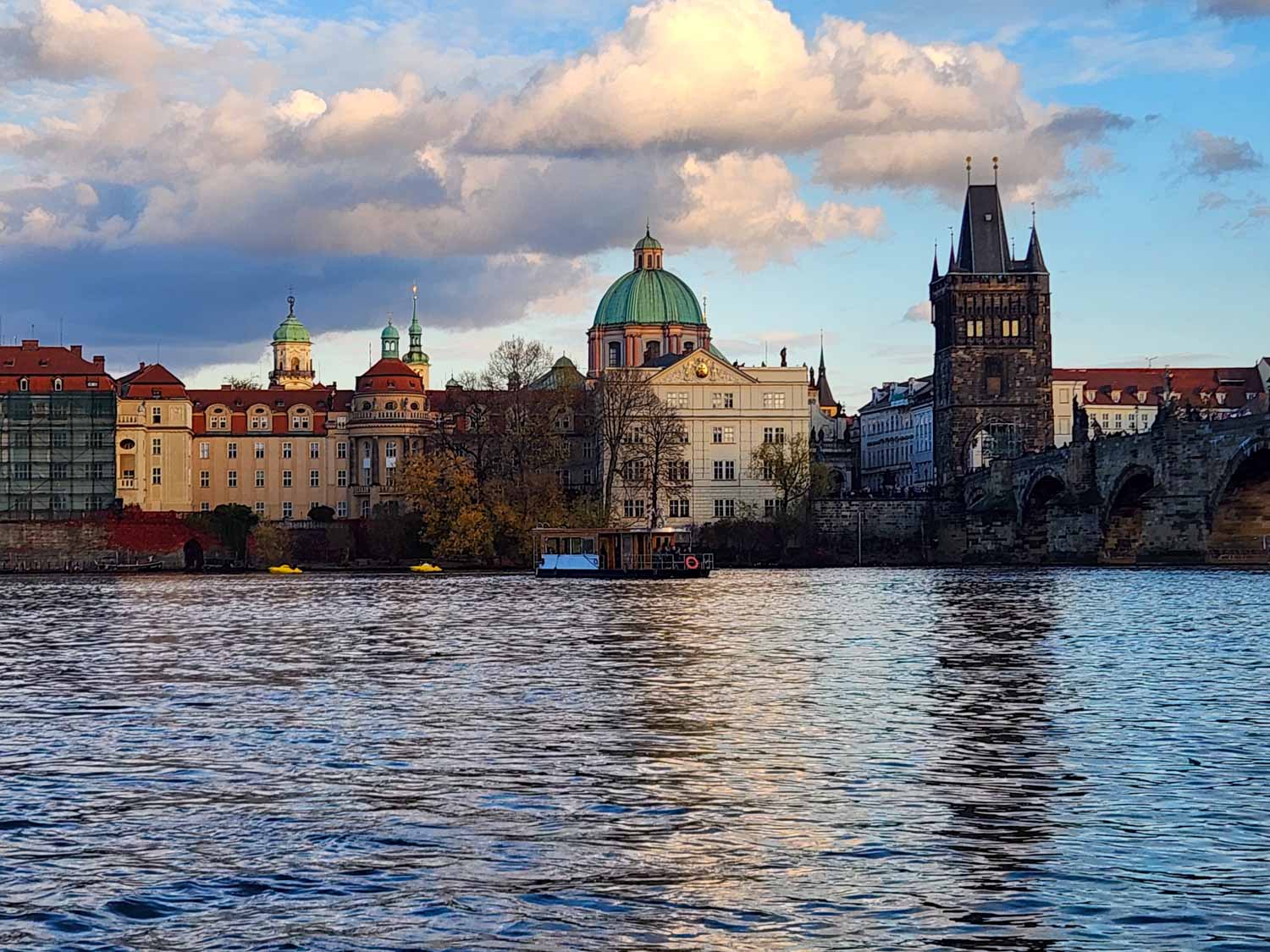 View of buildings along the river bank, a small wooden cruise boat and Charles Bridge around sunset during a river cruise in Prague with kids