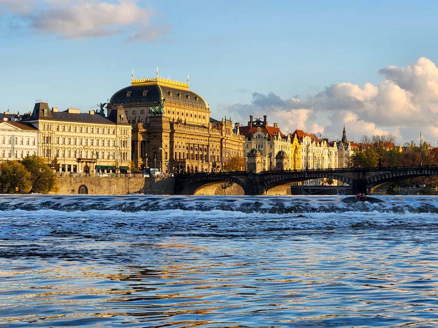 View to the grand buildings on the river bank, seen across a small weir on the Vltava - a river cruise is one of the best things to do in Prague with kids