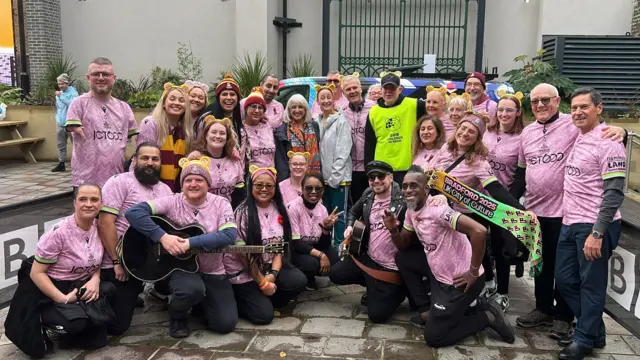 Colson Smith with members of the BBC Radio Leeds Bantam of the Opera choir gathered together in a courtyard who are wearing pink branded T-shirts and some of them are wearing Pudsey ears.