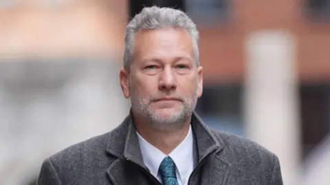 PA Media A grey-heaired man with a grey beard wearing a grey coat, blue shirt and a blue tie looking at the camera while walking away from the Old Bailey