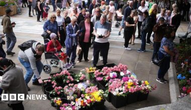 Scores of people crowd around pots of flowers at a outdoor vigil to mark a year since the 2024 attack. The flowers are mainly pink, white and yellow.