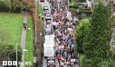 A drone shot of a protest held in Crowborough, East Sussex on 16 November over plans to house asylum seekers at an army site in the town. Hundreds of people can be seen walking on the streets with flags and banners. Cars are driving alongside the walkers.