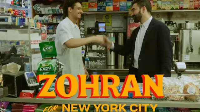 Zohran Mamdani shakes hands with a man in a white shirt in a deli with yellow lettering 'Zohran for New York City' in yellow lettering