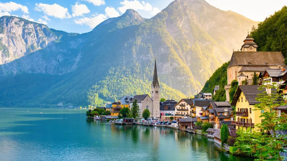Amidst towering mountains, charming houses imbue Hallstatt Austria with vibrant colors along the lakeside. The golden sunlight dances on calm waters, creating a picturesque reflection at dusk.