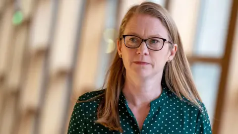 PA Media Social Justice Secretary Shirley-Anne Somerville has long reddish fair hair. She is wearing glasses and a green blouse with white dots. She is walking along a sunlit corridor.