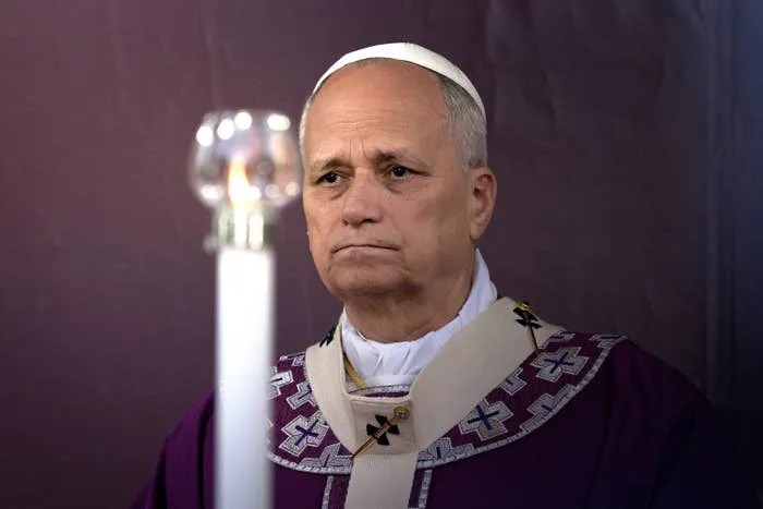 A person dressed in religious attire, wearing a mitre, stands solemnly behind a lit candle
