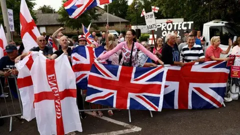 Reuters A group of people, some carrying large union jack and England flags. Some have placards with "Protect our..." and "Stop the boats" written on them. There are trees and a building behind them.