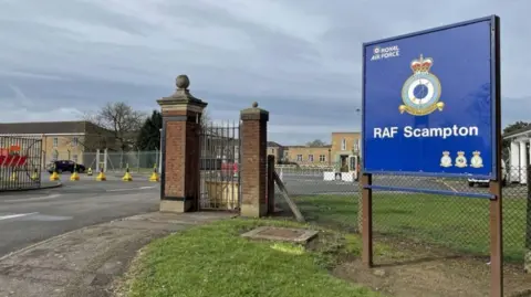 PA Media A blue RAF Scampton sign outside the former air base. Some of the brick buildings can be seen behind the fence. There is grass and a white car parked in the distance.